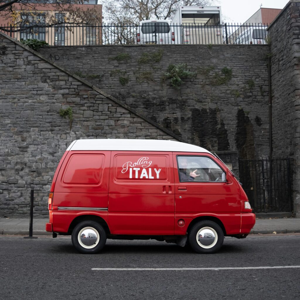 Side view of a red camper van with white roof driving past a stone wall in Bristol, UK.